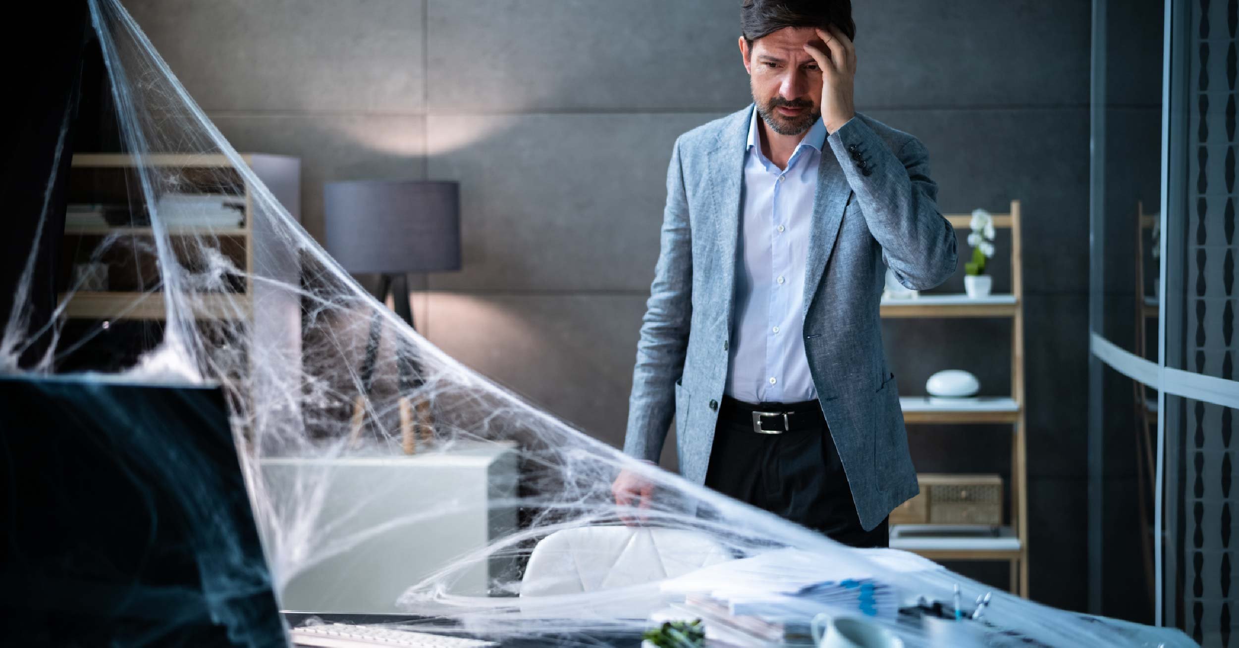 A male employee regards a computer and keyboard covered with cobwebs with dismay.