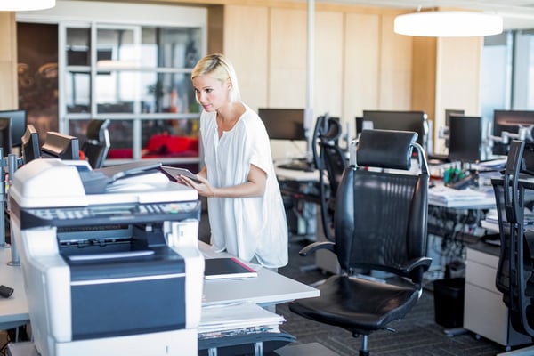 Image description: Woman holding a tablet leans over a multi-function printer. She is in a computer lab.