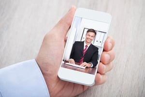 Image description: hand holding a smartphone depicting a mobile conference call with a businessman in a black blazer and red tie. 