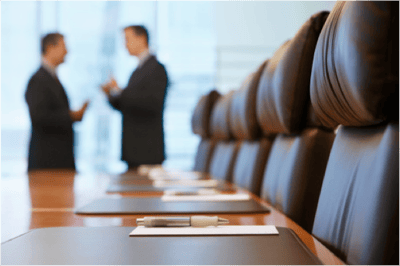 Image description: Boardroom with chairs, folders, pens and two blurred individuals standing in the foreground.